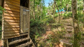 Una cabaña de madera con puerta y un pequeño porche, rodeada de un bosque frondoso y un sendero visible.