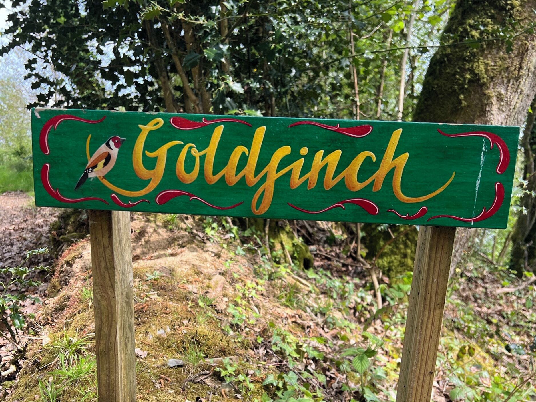 A wooden sign with the name 'Goldfinch' and a bird illustration, set against a natural backdrop.