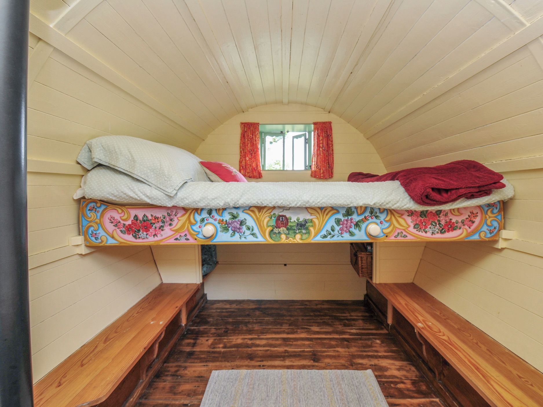 Quarto de sótão pequeno e aconchegante, com uma cama de solteiro, bancos de madeira e janela com cortinas.