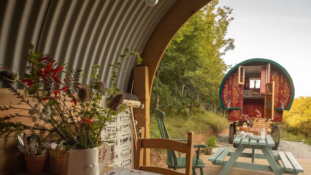 Un coin repas avec une table dressée pour deux personnes, une plante en pot et une vue sur un bâtiment rouge, visible à travers une arche.