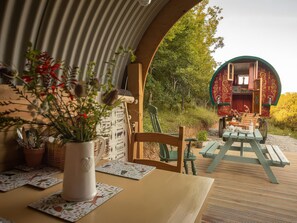 Un coin repas avec une table dressée pour deux personnes, une plante en pot et une vue sur un bâtiment rouge, visible à travers une arche.