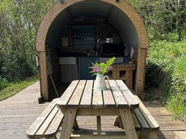 A wooden archway leading to a kitchen with cabinets and a sink, a wooden picnic table with a vase of flowers, and a wooden deck.