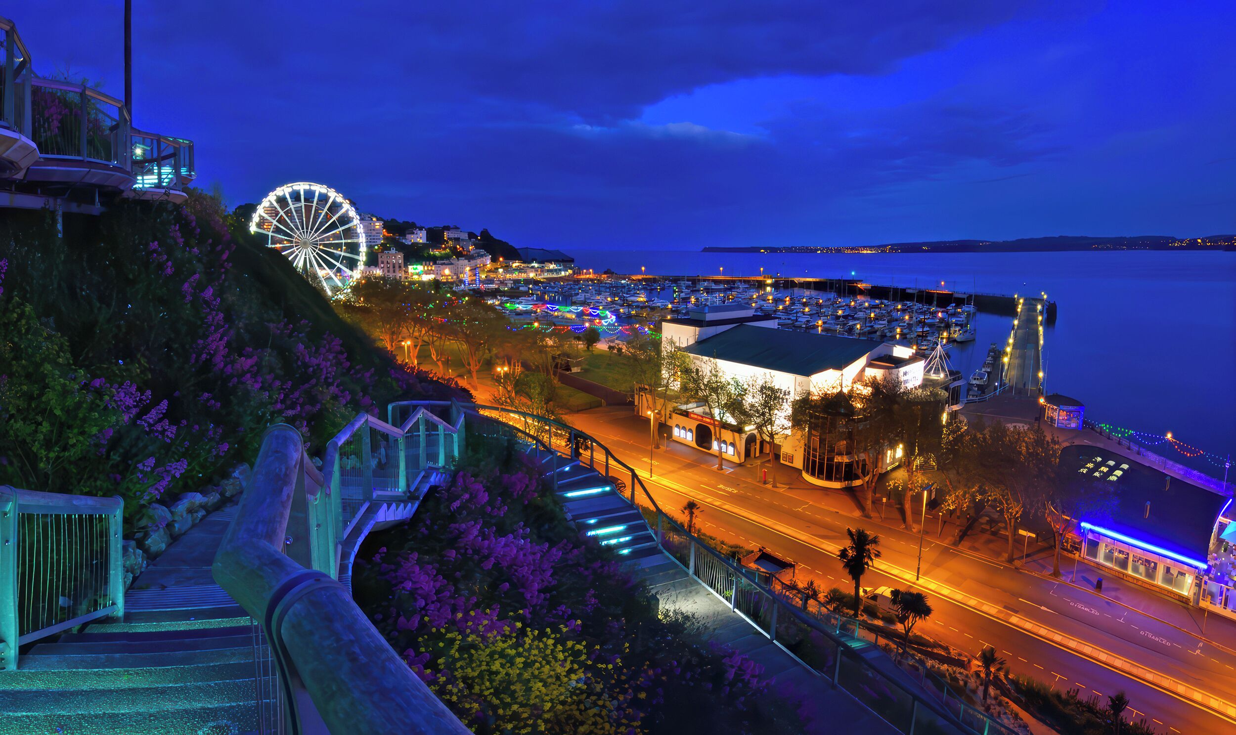 A coastal cityscape at night with a prominent Ferris wheel, a marina, and a brightly lit staircase leading down to the waterfront.