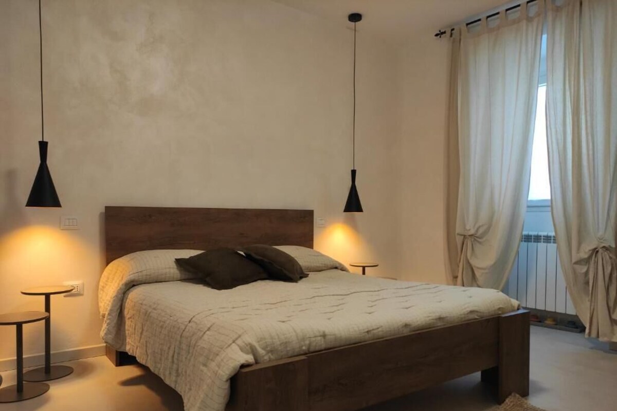 A bedroom with a wooden bed, two bedside lamps, a small table, and a view of the outdoors through curtains.