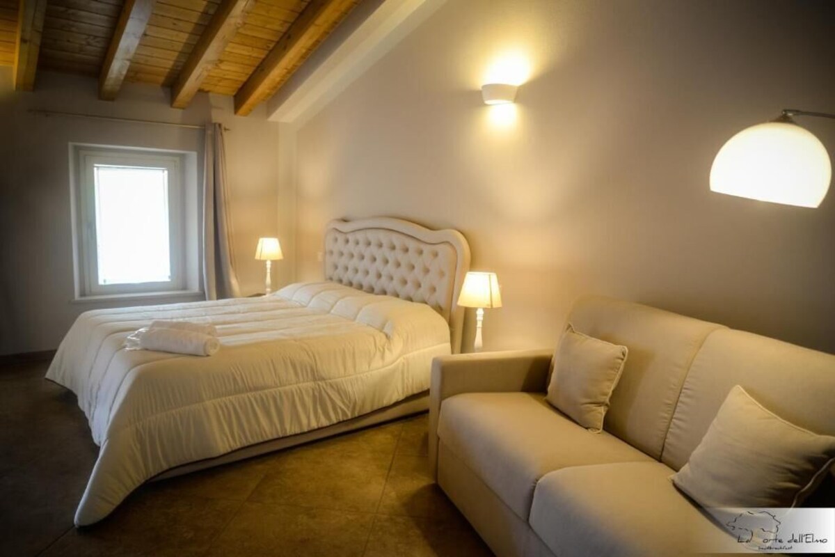 A bedroom with a double bed, a sofa, two bedside lamps, and a window with curtains.