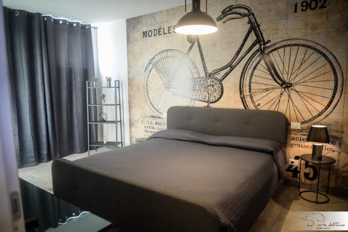 A bedroom with a large bed, a wall-mounted bicycle illustration, a small side table with a lamp, and a shelf with decorative items.