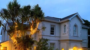 Un edificio ampio su due piani, con una facciata simmetrica, numerose finestre e un giardino curato con palme e piante fiorite.
