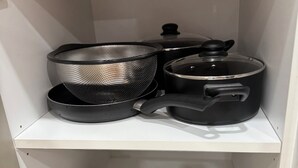 Ett köksskåp med ett metallstrains, en svart kastrull med lock och två stekpannor.