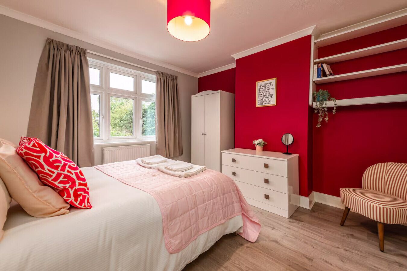 A bedroom with a bed, a wardrobe, a chair, and a shelf with books and a plant.