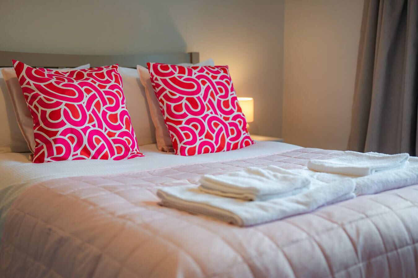 A neatly made bed with pink and white patterned pillows, a folded white towel, and a bedside lamp.
