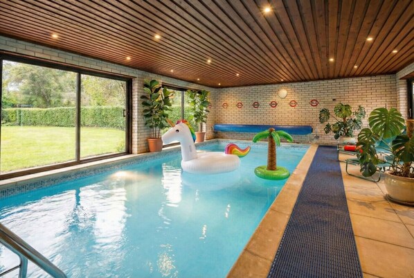 An indoor swimming pool with a wooden ceiling, a white inflatable unicorn float, and potted plants.