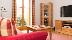 A living room with a red sofa, a wooden cabinet, a television, and a window with curtains. - Landhaus Albrecht - Holiday Apartment 01 - with 2 terraces and fireplace (50 m²) (Mittenwald)