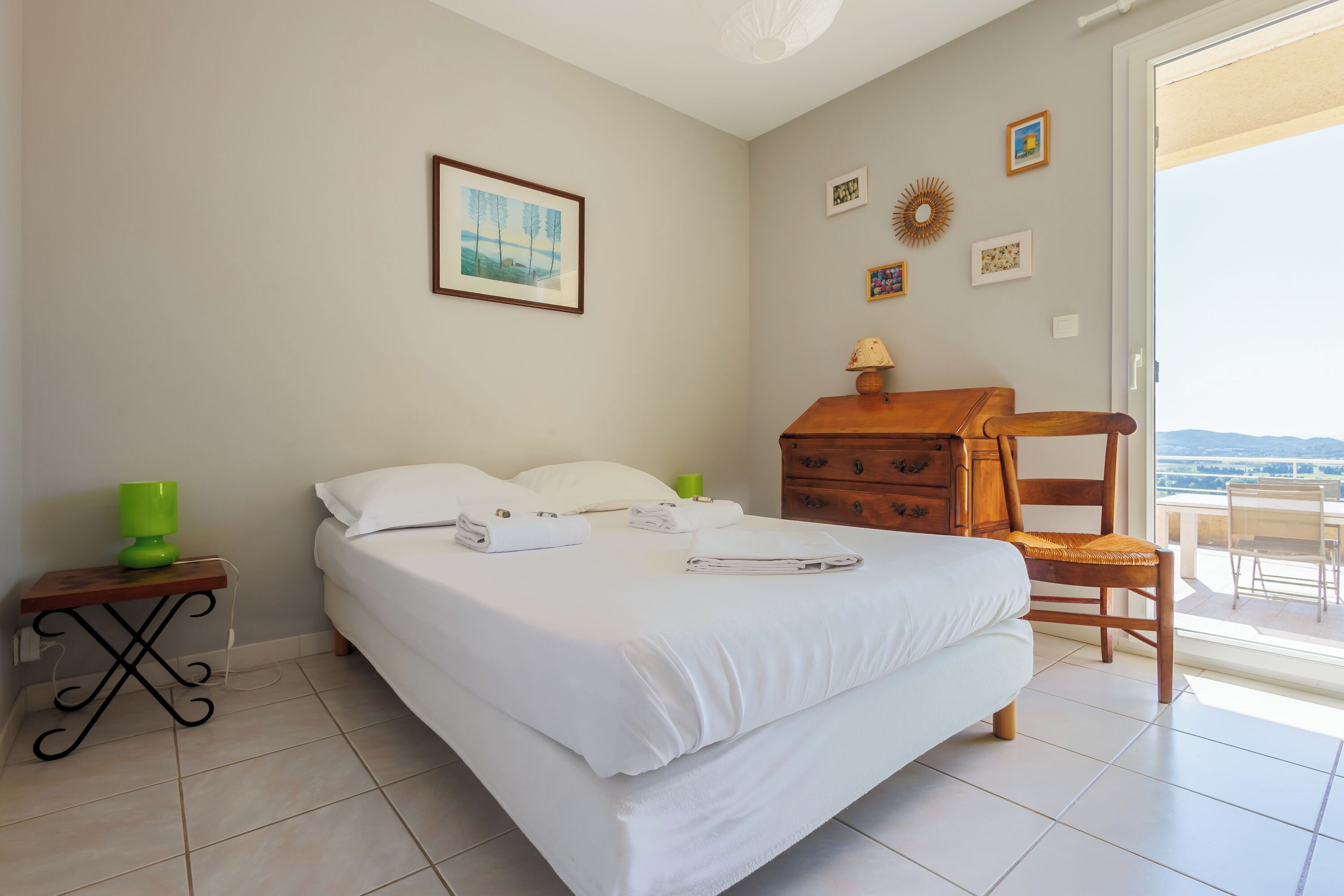A neatly made bed with white linens, a wooden nightstand with a green lamp, and a wooden dresser with a lamp.