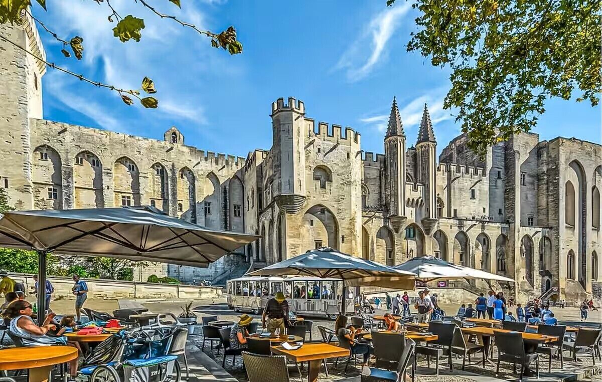 Ein Straßencafé mit Tischen und Sonnenschirmen vor einem historischen Steinhaus mit Bogenfenstern und Türmen.