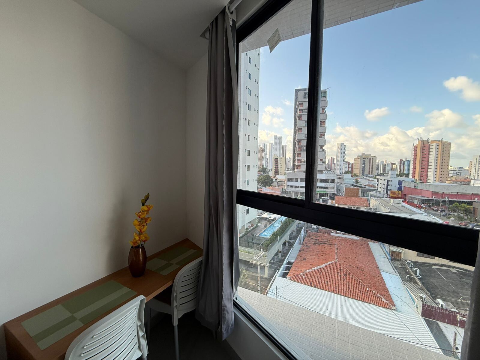 A room with a view of a cityscape, a desk with a chair, and a vase with flowers.