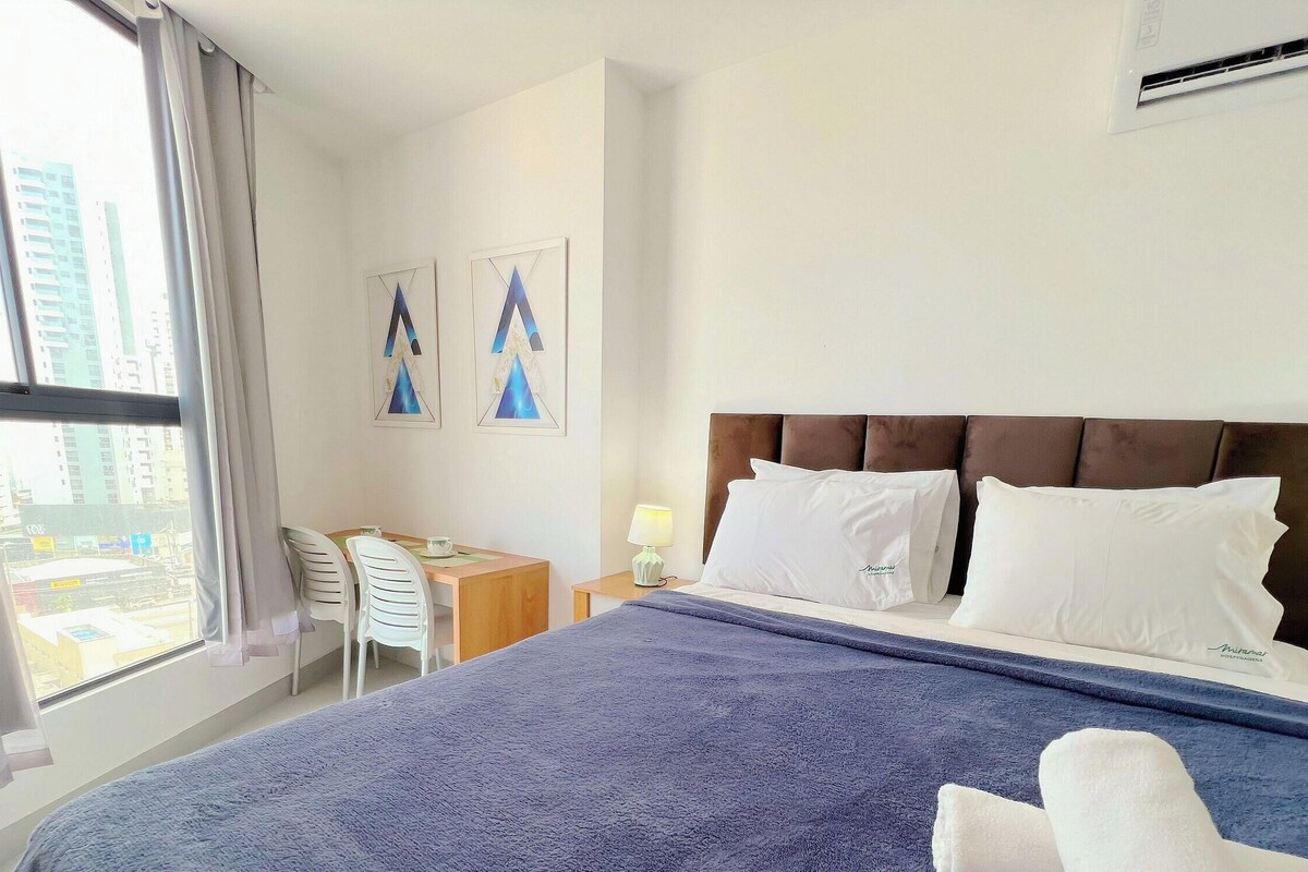Ein Hotelzimmer mit einem Bett, einem Schreibtisch, zwei Stühlen, einer Lampe und einem Fenster mit Blick auf die Stadt.