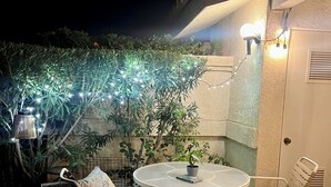Eine Terrasse im Freien mit einem runden Tisch und zwei Stühlen, umgeben von Blumentöpfen und Lichterketten.