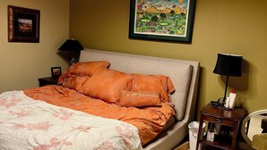 Ein ordentlich bezogenes Bett mit orangefarbener und weißer Bettwäsche, ein gerahmtes Landschaftsbild darüber und ein kleiner Nachttisch mit Lampe und Flasche.
