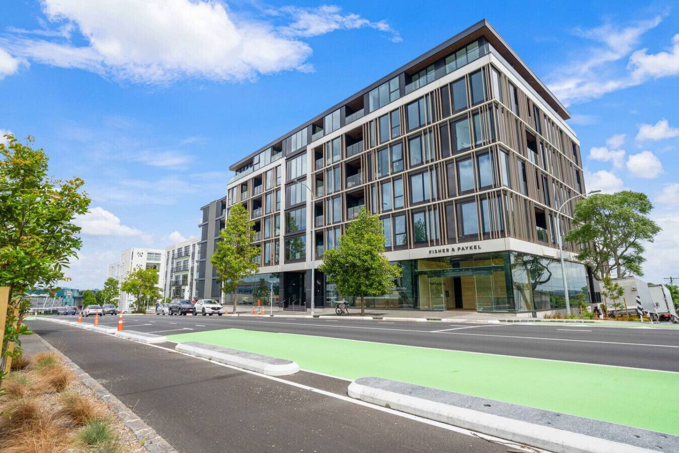 A modern building with a glass facade, labeled "FISHER & PAYKEL," featuring a green bike lane and trees in the foreground.