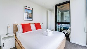 Et moderne hotelværelse med en stor seng, et natbord, et billede i ramme på væggen og udsigt over byen gennem et stort vindue.
