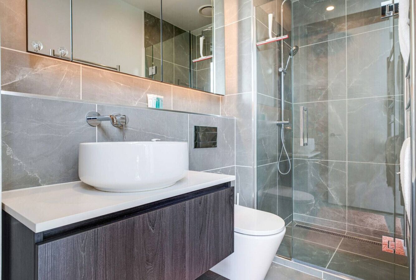 Un baño moderno con un lavamanos grande de color blanco, tocador de madera oscura y cabina de ducha con paredes de vidrio.