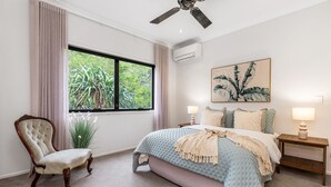 Ein Schlafzimmer mit großem Fenster, Deckenventilator, Bett mit blauer Steudecke, Stuhl und Nachttisch mit Lampe.