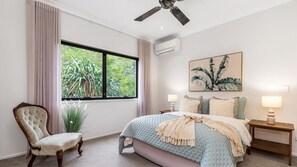 Ein Schlafzimmer mit großem Fenster, Deckenventilator, Bett mit blauer Steudecke, Stuhl und Nachttisch mit Lampe.