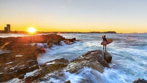 Eine Person steht auf einem felsigen Vorsprung und hält ein Surfbrett, im Hintergrund schimmern Stadtansicht und Sonnenuntergang.