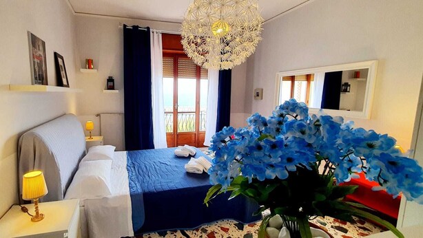 Ett sovrum med en säng, sänglampor, en kristallkrona, ett fönster med gardiner och en vas med blå blommor.