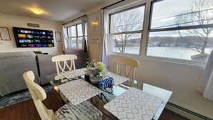 A dining area with a glass table, white chairs, a floral centerpiece, and a TV displaying various channels. - 1 Bed 1 bath Lakefront Apartment in Lake Hopatcong, NJ w/ lake access and kayaks (Jefferson)
