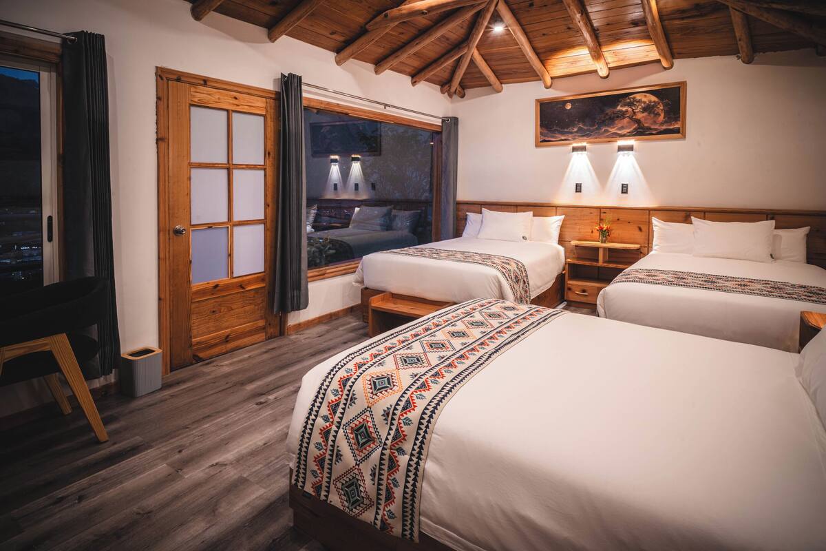 Habitación de hotel con dos camas, suelo de madera, un ventanal grande y techo de madera.