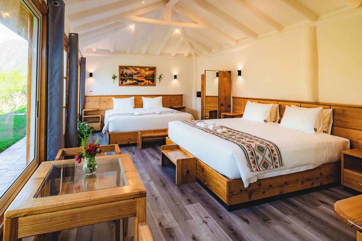 Un dormitorio amplio con una cama grande, una cama más pequeña, un banco de madera, una mesa de vidrio con un florero y vistas al exterior.