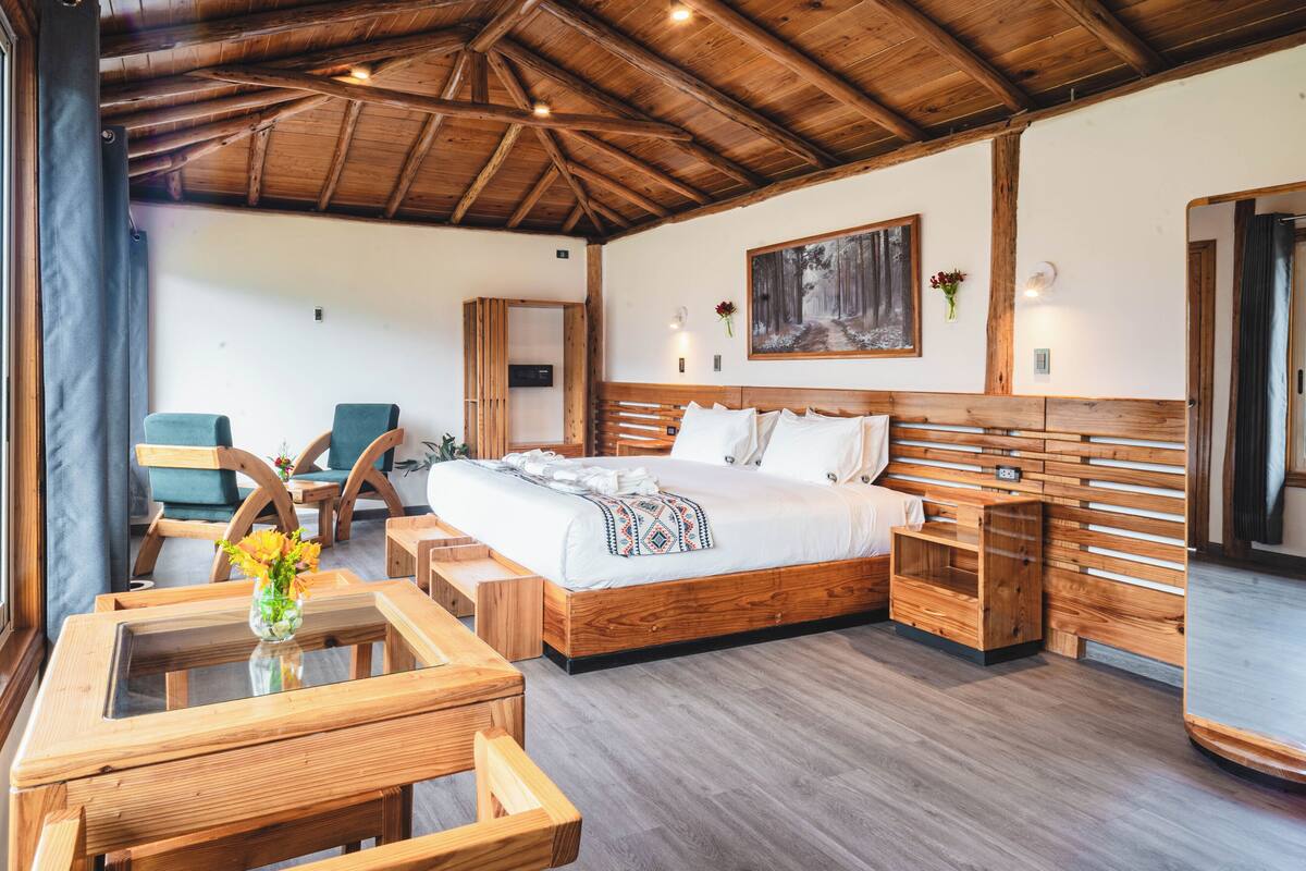 Una cama con estructura de madera y una colcha blanca, un cuadro en la pared y un banco de madera con un jarrón de flores.