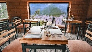 Een restaurant met een rustieke bakstenen muur en grote ramen met uitzicht op de bergen. Tafels zijn gedekt met wijnglazen en menukaarten.
