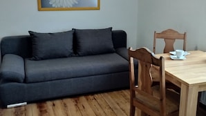 A room with a grey sofa, a wooden dining table with chairs, and a framed flower painting on the wall. - Vacation/ mechanic apartment Nikki (Argenthal)