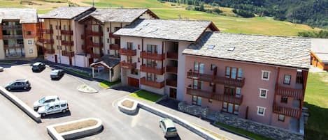 Un edificio residencial de varios pisos con balcones, un estacionamiento y montañas al fondo.