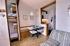 Ein kleines Zimmer mit Etagenbett, Esstisch mit Stühlen, einer Couch und einer Küchenzeile.