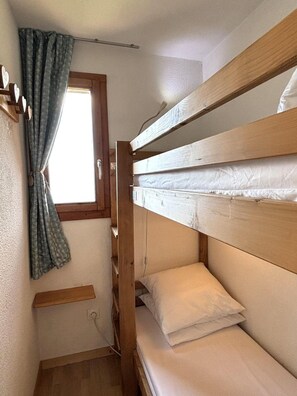 Une petite pièce étroite, avec un lit superposé, une fenêtre avec des rideaux et une étagère en bois.