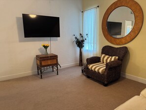 Ein Wohnzimmer mit einem wandmontierten Flachbildfernseher, einem Korbstuhl mit gestreifter Sitzkissen, einem Holztisch mit Blumenvase und einer hohen Vase mit dekorativen Ästen.