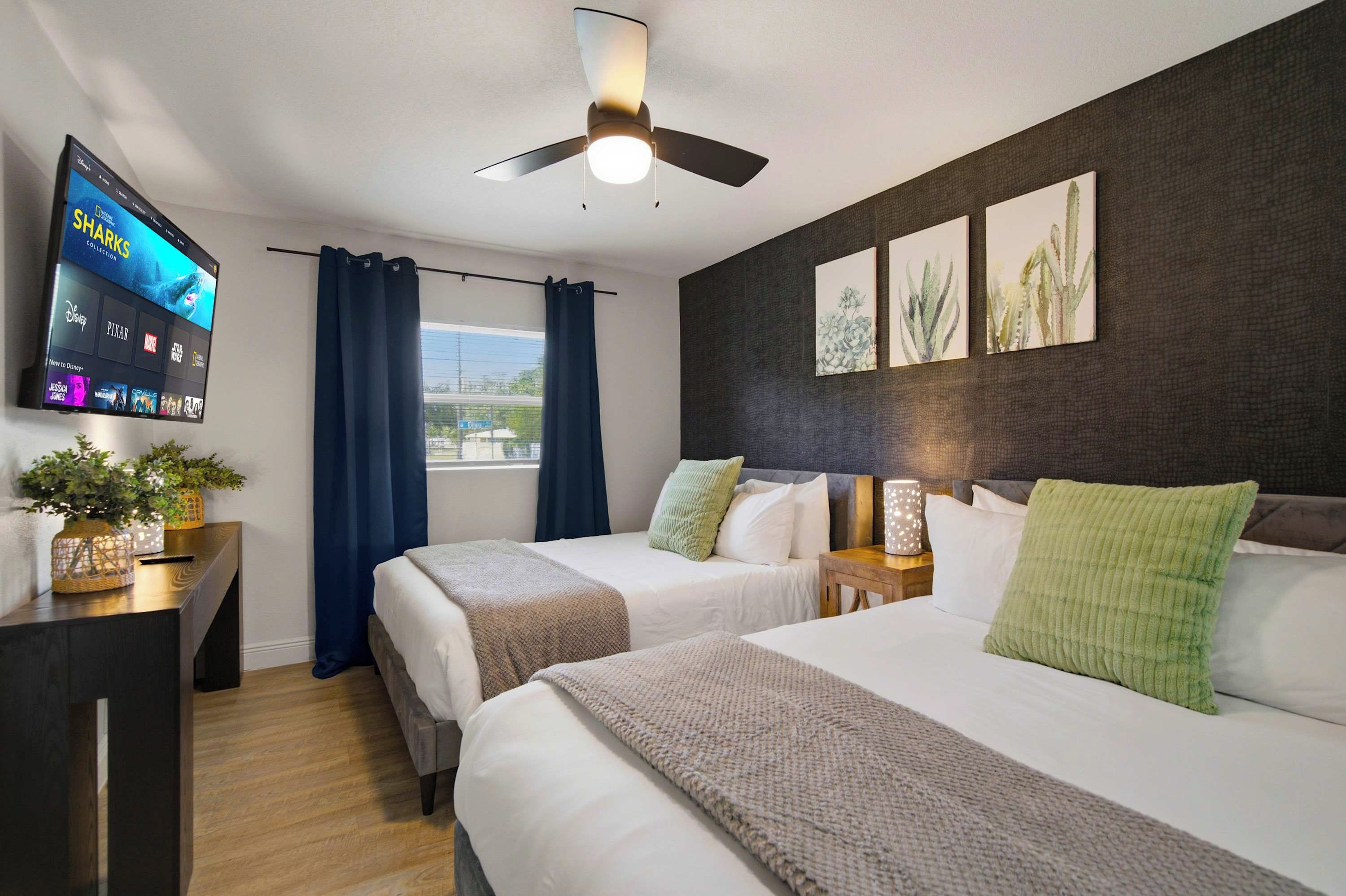 A hotel room with two beds, a flat-screen TV, a ceiling fan, and artwork on the wall.