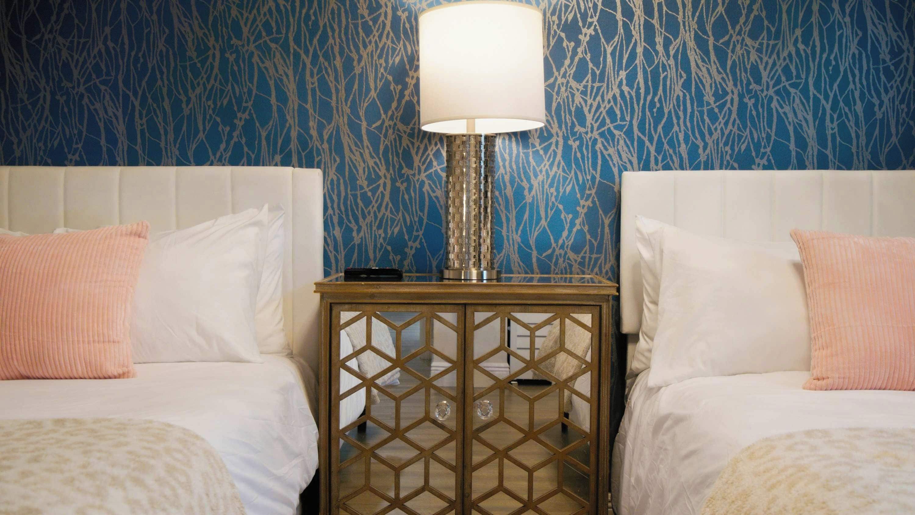 A hotel room with two beds, a nightstand with a lamp, and a patterned wall.
