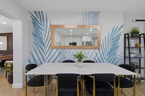 A dining area with a white table, black chairs with gold legs, a large mirror, and a wall with a blue leaf pattern. - Tampa Backyard Oasis | Pool • Golf • Glam Room (Tampa)