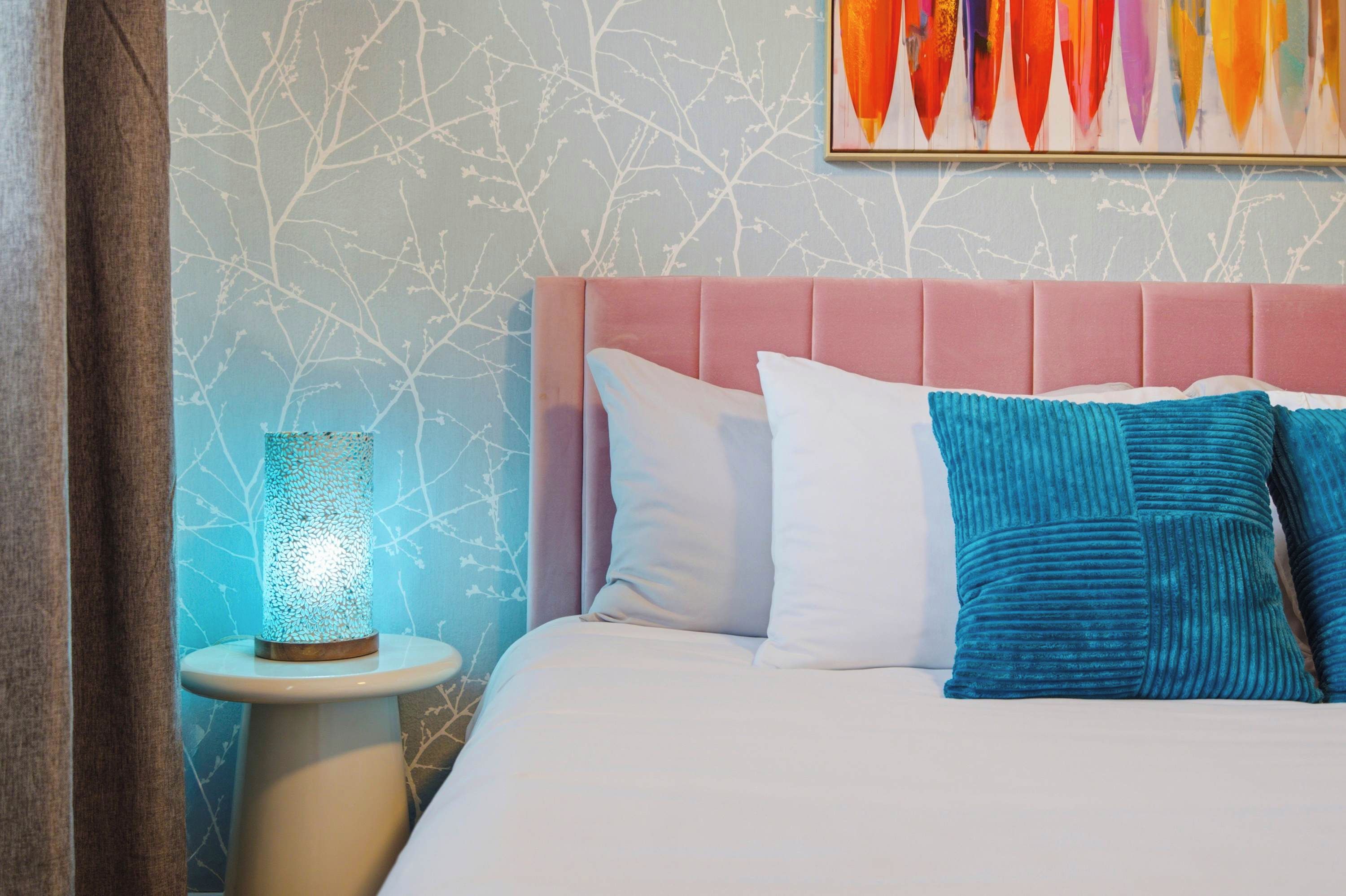 Une chambre à coucher avec un lit, des oreillers, une table de chevet, une lampe et un mur orné d’un motif représentant une branche d’arbre.