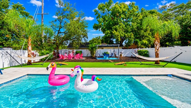 A swimming pool with inflatable swan and flamingo toys, surrounded by a grassy lawn and trees.