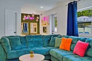 A teal-colored sectional sofa with multiple cushions, a small round table with a plant, and a neon sign in the background. - Tampa Backyard Oasis | Pool • Golf • Glam Room (Tampa)