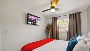 A hotel room with a bed, a ceiling fan, a TV mounted on the wall, and a window with curtains.