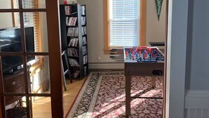 Una habitación con una mesa de futbolín, una estantería, una ventana con persianas y una alfombra estampada.