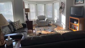 Una sala de estar con un sofá de cuero oscuro, un sillón a cuadros, una mesa de centro con libros y una lámpara, y una estantería con un televisor.