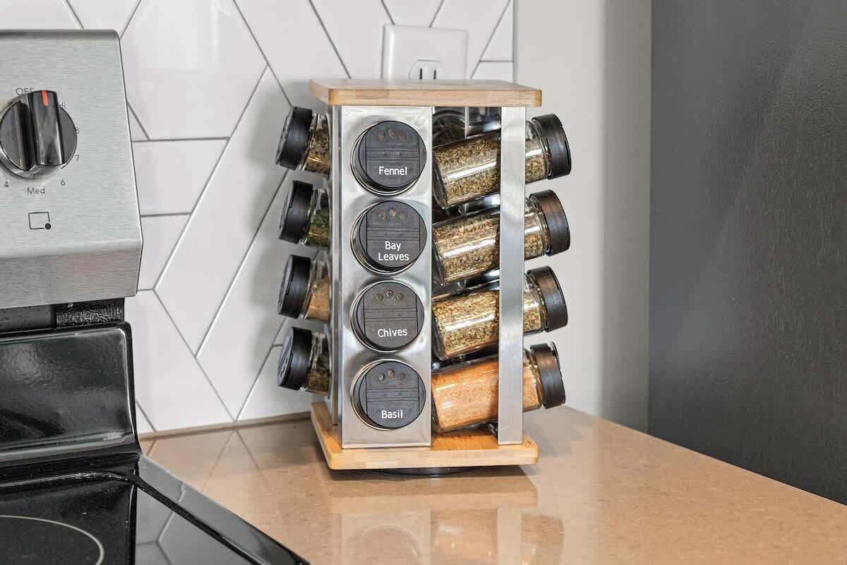 A kitchen with a stainless steel range hood, a wooden spice rack with labeled compartments for fennel, bay leaves, chives, and basil, and a tiled backsplash.