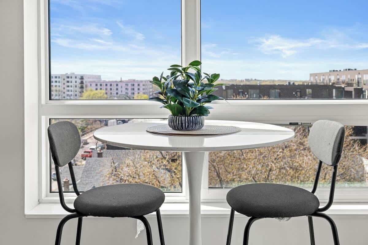 Una habitación con una mesa redonda y dos sillas, una planta en maceta sobre la mesa, y vistas de edificios a través de un ventanal grande.